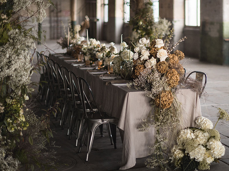 Long banquet table with black metal chairs, grey tablecloths and abundant arrangements of white and dried flowers in a spacious industrial-style room.