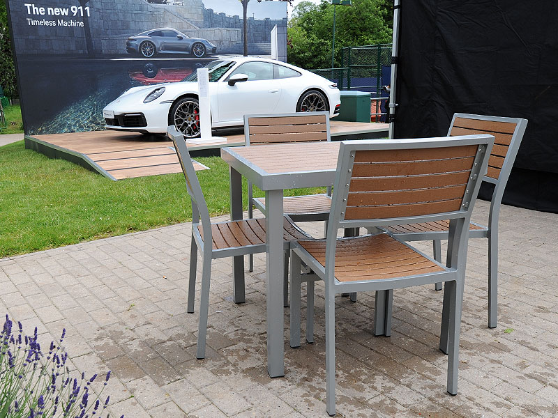 Outdoor table with four wood-panelled chairs on paving, with a white sports car displayed on a raised platform beneath a banner reading “The new 911 – Timeless Machine”.