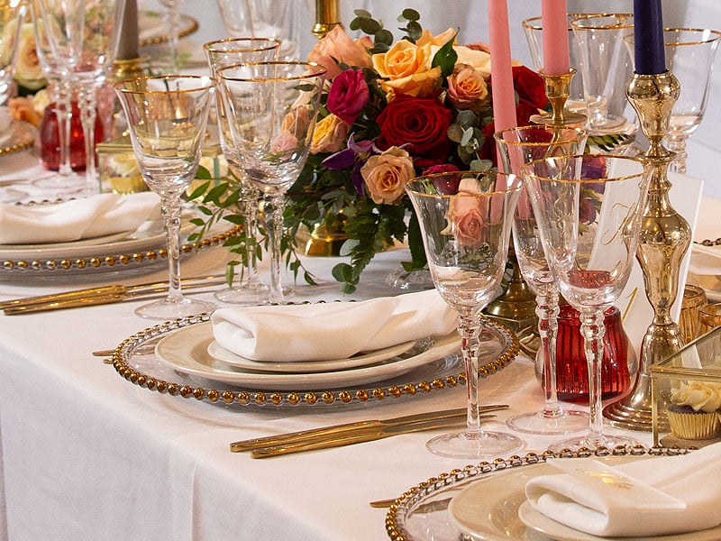Elegant banquet table setting with gold-rimmed glassware, white plates and napkins, gold cutlery, colourful roses and tall pink and purple candles.
