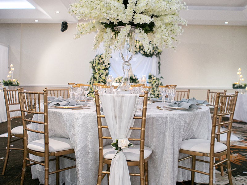 Round banquet table with a white textured cloth, grey napkins and gold chairs, centred beneath a tall glass vase overflowing with white flowers.