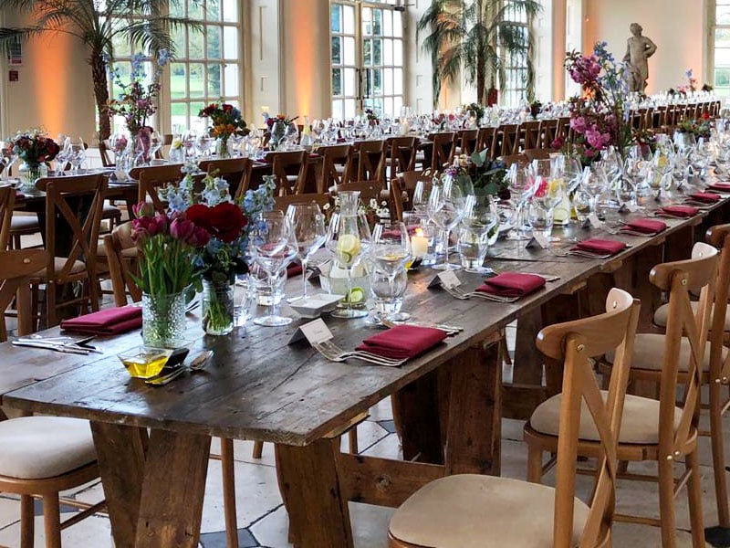 Long rustic wooden banquet tables set with glassware, burgundy napkins and colourful floral arrangements in a bright hall with tall windows, palm trees and a statue.