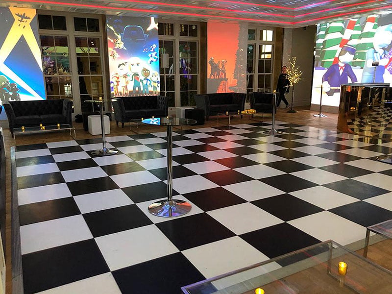 Modern event space with a large black-and-white chequered floor, black lounge seating, tall glass tables and colourful projected artwork covering the walls.