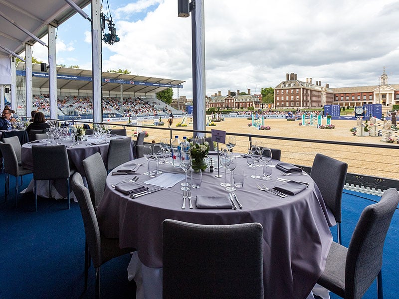 Round dining tables set with grey tablecloths, glassware and cutlery overlook an outdoor equestrian arena with colourful show-jumping fences and grandstands.