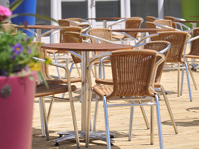 Empty outdoor café seating with wicker chairs and wooden-topped tables on a sunlit deck, with a blurred planter of colourful flowers in the foreground.