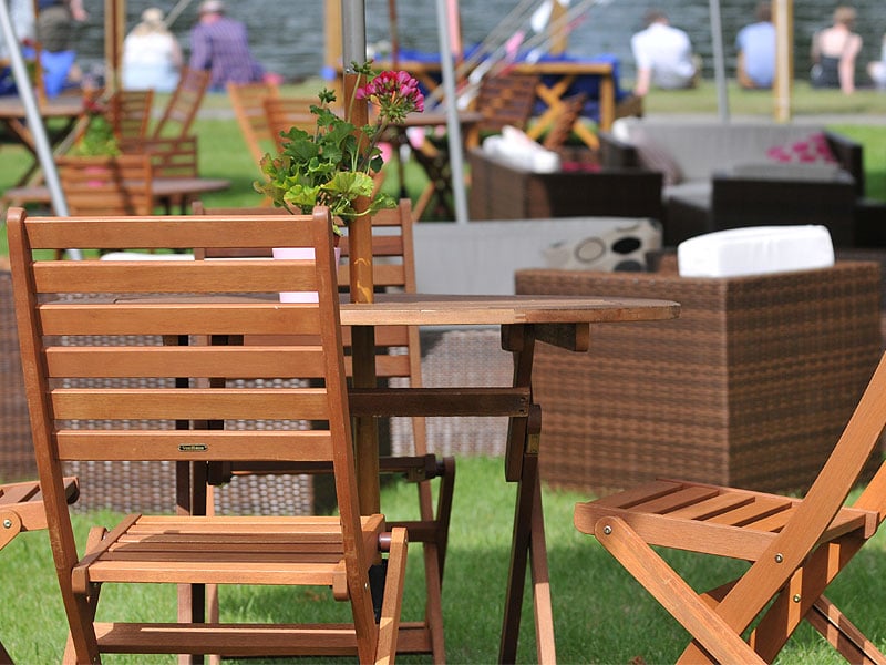 Wooden folding chairs and tables with a vase of pink flowers stand on grass beside wicker outdoor seating, with people blurred in the background.