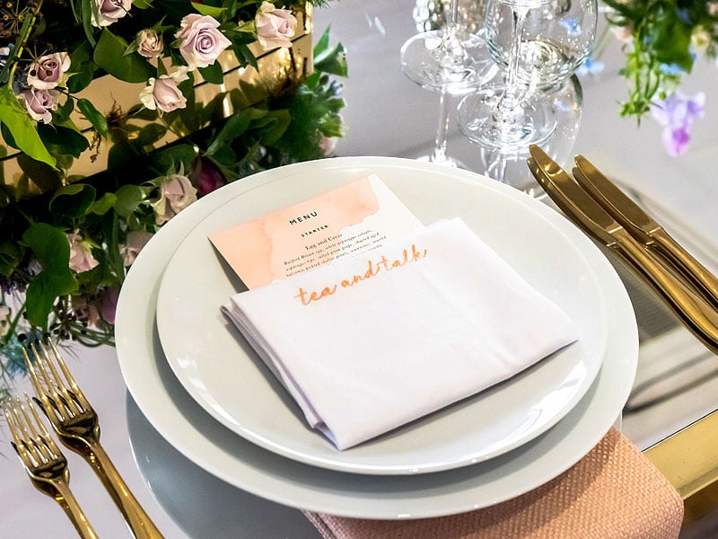 Elegant place setting with stacked white plates, a folded napkin labelled “teamandtalk”, a printed menu, gold cutlery, glassware and pale pink roses.