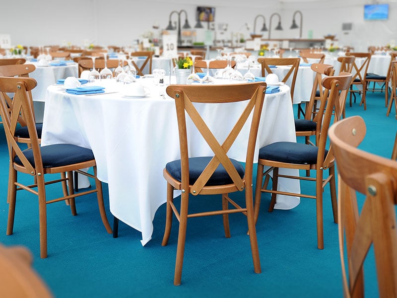 Event dining area with round tables covered in white cloths, wooden cross-back chairs, blue napkins and glassware on a turquoise carpet.
