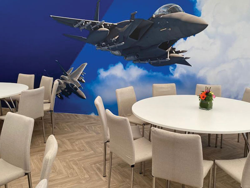 Round white tables with cream upholstered chairs in front of a wall mural showing two military fighter jets flying through clouds.