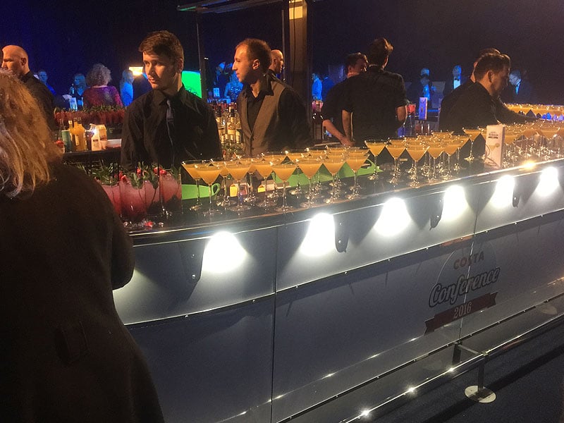Busy event bar with staff behind an illuminated counter lined with rows of cocktails, with “Costa Conference 2016” branding on the front.