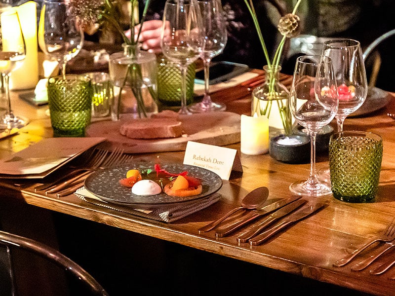 Candlelit dining table set with wine glasses, green textured tumblers, cutlery, small flower vases and a plated appetiser beside a place card.
