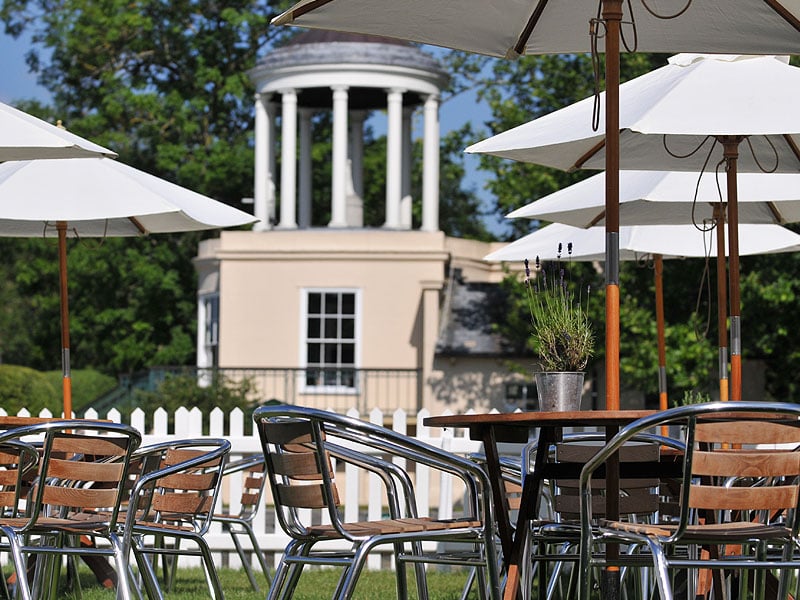 Outdoor café tables and metal chairs beneath white parasols, with a potted lavender plant and a columned pavilion in the background.