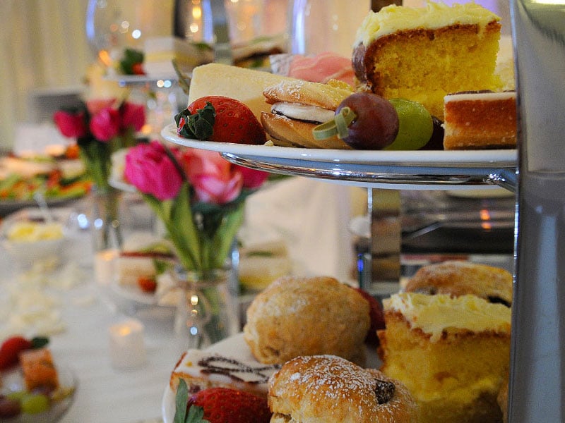 Tiered afternoon tea stand filled with cakes, scones, macarons, strawberries and grapes, with pink flowers and other refreshments blurred in the background.