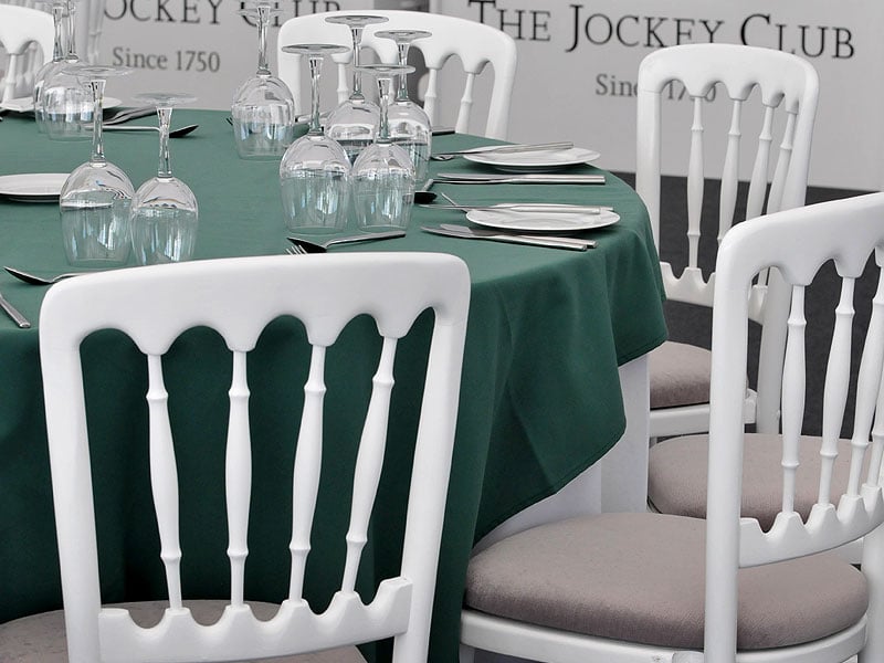 Round dining table with a dark green tablecloth, white chairs, plates, cutlery and upside-down wine glasses, set against a backdrop reading “The Jockey Club”.