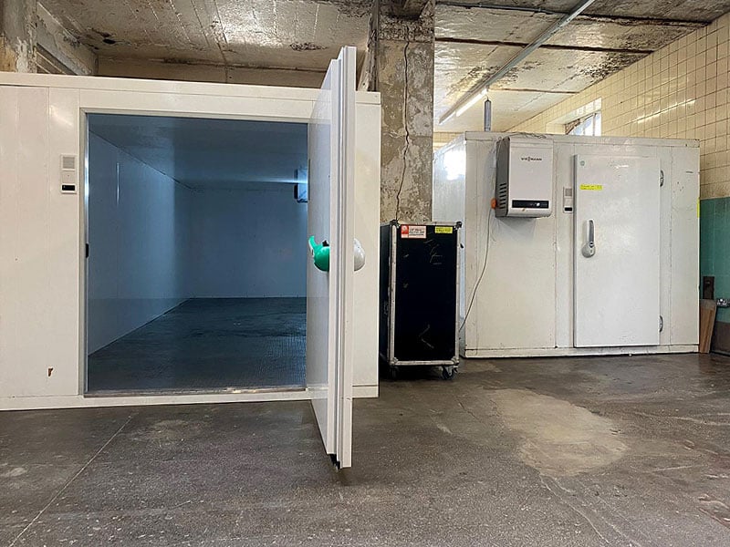 Two large white cold-storage units stand in an industrial room, with the nearer unit’s thick door open to reveal an empty interior.