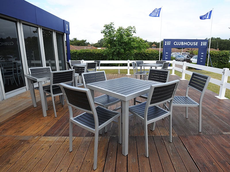 Grey outdoor tables and chairs arranged on a wooden clubhouse terrace, overlooking a fenced entrance with blue flags.