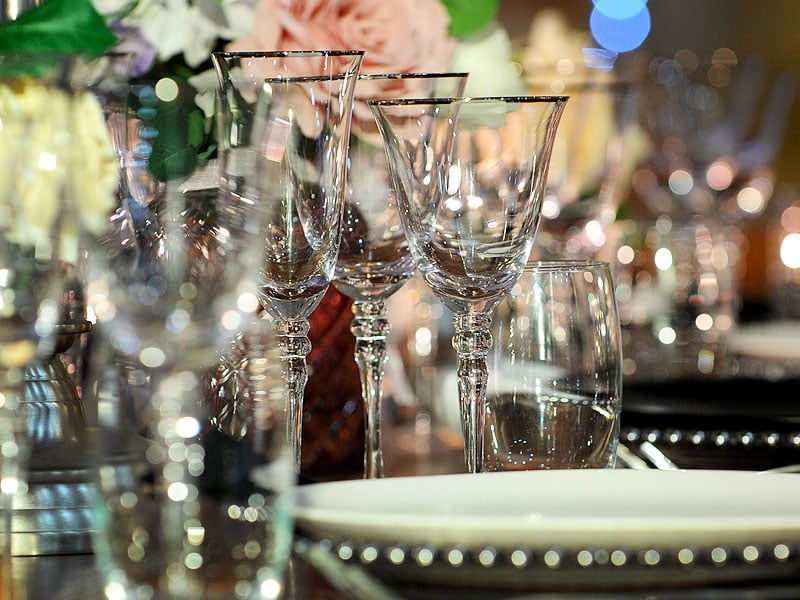 Close-up of clear wine glasses and tumblers arranged on a formally set dining table, with soft reflections and blurred flowers in the background.