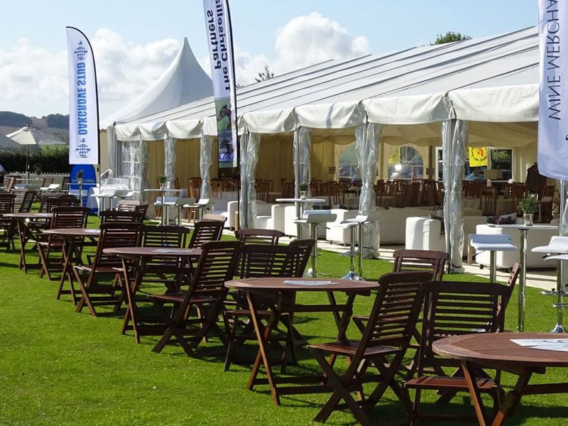Outdoor seating with wooden tables and chairs on grass beside a large white marquee, with white lounge furniture and tall event banners.
