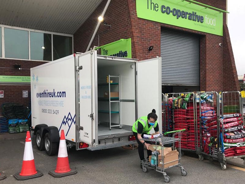 A worker in a high-visibility vest unloads boxes from an open event-hire trailer beside wheeled racks of bagged goods outside a Co-operative Food building.