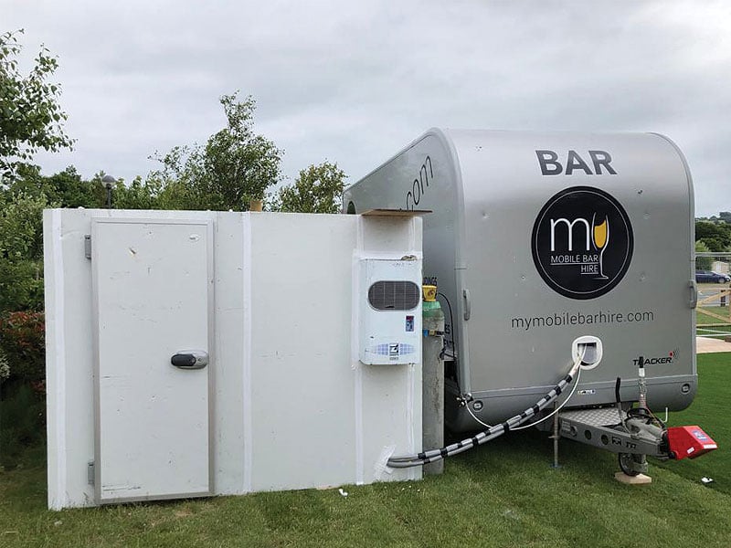 Silver mobile bar trailer parked on grass beside a white temporary enclosure, with “BAR” and “mymobilebarhire.com” visible on the trailer.