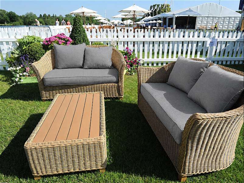 Outdoor wicker seating area on grass with two grey-cushioned sofas and a low rectangular table, bordered by flowers and a white picket fence.