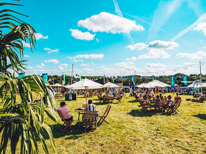 Outdoor festival seating area on a grassy field with wooden chairs, tables and white parasols, surrounded by groups of people beneath a bright blue sky with scattered clouds.