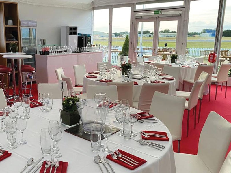 Bright event dining area with round tables set with glassware, cutlery and red napkins, surrounded by white chairs on a red carpet, with large windows overlooking a grassy outdoor area.