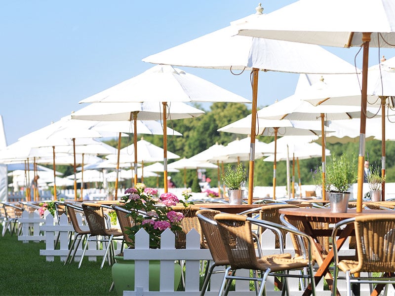 Outdoor café seating with wicker chairs and wooden tables beneath rows of large white parasols, surrounded by potted plants and colourful flowers.