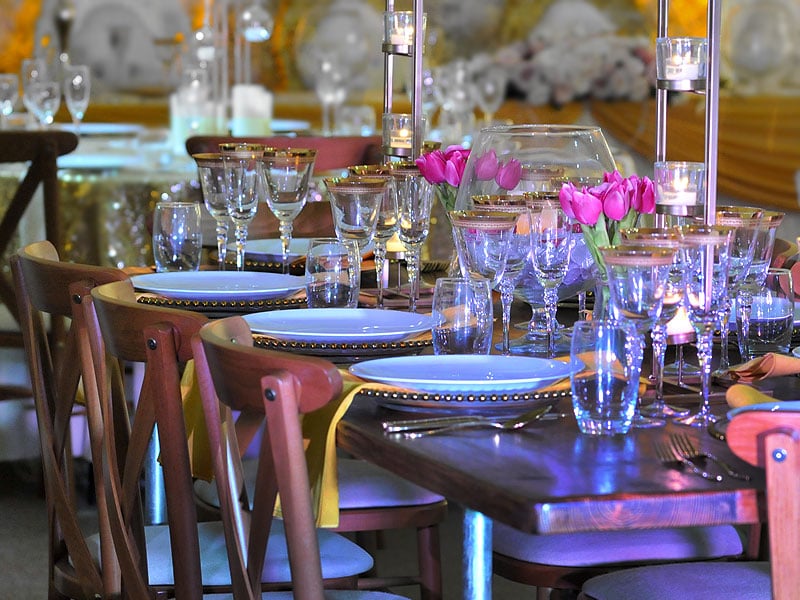 Formal dining table set with white plates, gold-rimmed glassware, tall candle holders and bright pink tulips, surrounded by wooden chairs.