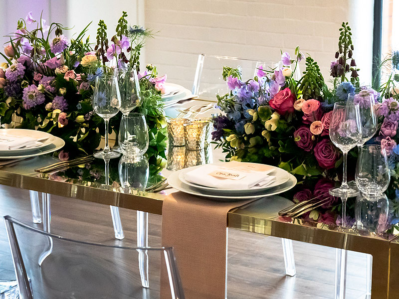 Gold-framed dining table set with white plates, glassware and blush-pink runners, surrounded by abundant purple, pink and blue floral arrangements and candle holders.