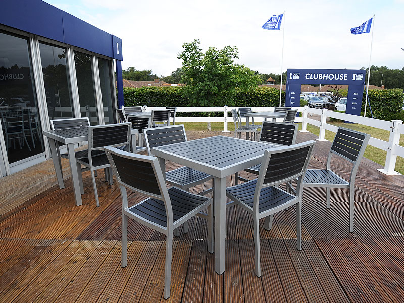 Grey outdoor tables and chairs arranged on a wooden clubhouse terrace, with a white railing, blue flags and a car park beyond.