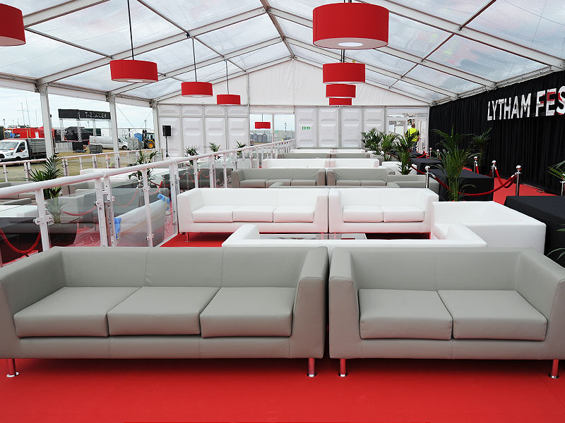 Event lounge inside a clear-roofed marquee, with rows of white sofas on red flooring, red pendant lights, potted plants and a roped-off stage area.