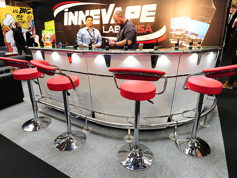 Curved illuminated bar with red cushioned stools, bottles and cups on the counter, and two men serving beneath a large “Inn Vape Lab USA” sign.