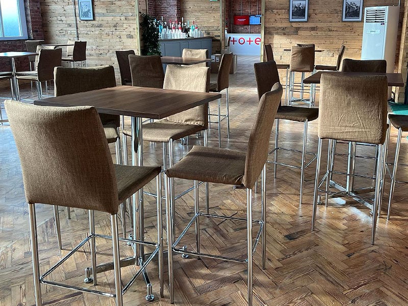 Café-style seating area with tall brown upholstered chairs around dark wooden tables on a parquet floor, against wood-panelled walls.