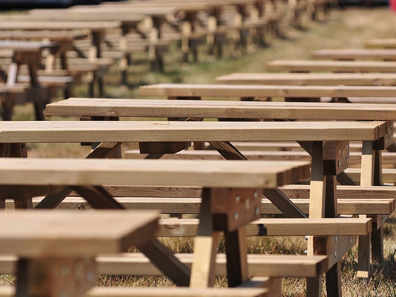 Rows of wooden picnic tables and benches arranged outdoors on grass, with the foreground in sharp focus and the background blurred.