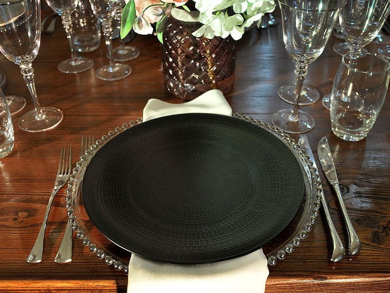 Formal table setting with a large black plate on a clear beaded charger, silver cutlery, folded white napkin, glassware and a floral centrepiece on a wooden table.