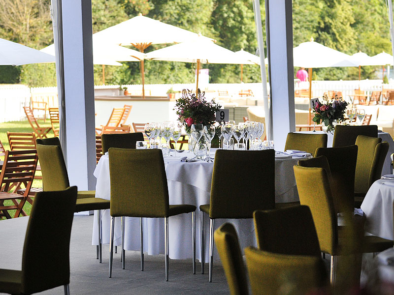 Indoor dining area with round tables, olive-green chairs, white tablecloths, glassware and floral centrepieces, overlooking a sunny terrace with wooden seating and large white parasols.
