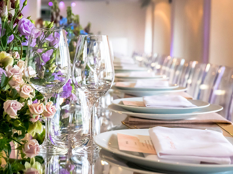 Elegant dining table set with white plates, folded napkins and wine glasses beside a floral arrangement of pale pink and purple flowers.