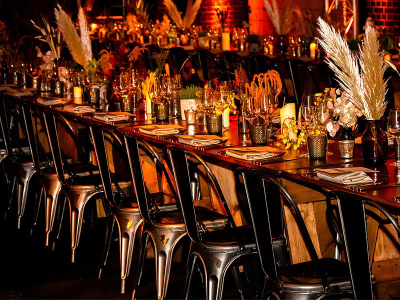 Long wooden dining tables set with plates, glassware, candles and dried floral arrangements, surrounded by metal chairs in warm orange lighting.