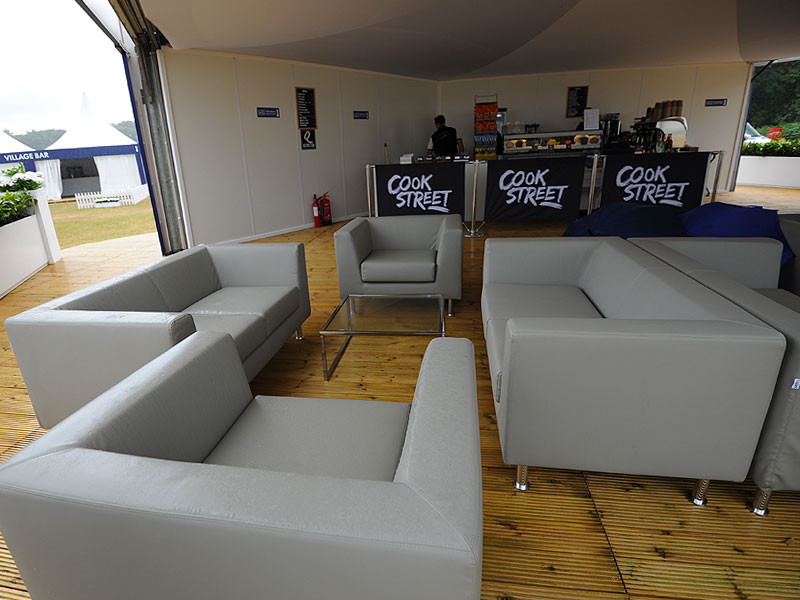 Grey sofas and armchairs arranged around a glass coffee table in a covered seating area, with a “Cook Street” café counter in the background.