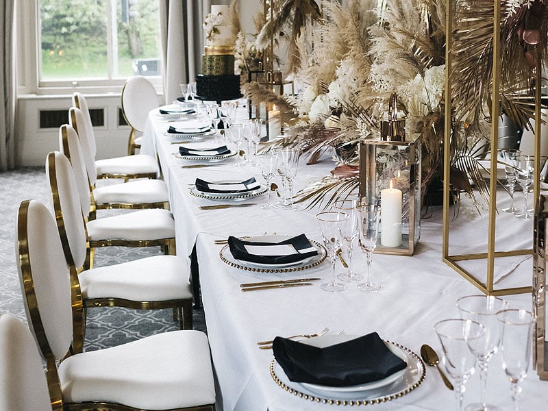 Elegantly set dining table with white chairs, black napkins, gold-toned cutlery, glassware, candles and tall arrangements of dried grasses and foliage.