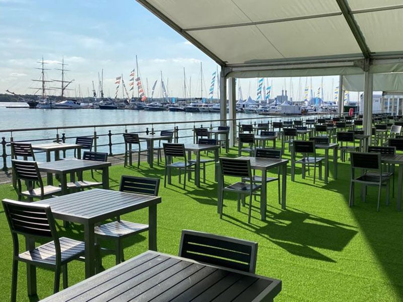 Empty covered terrace with black tables and chairs on artificial grass, overlooking a marina filled with yachts and colourful sails.