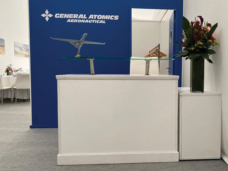 White reception counter with a glass top in front of a blue General Atomics Aeronautical display featuring an aircraft model, beside a tall floral arrangement.