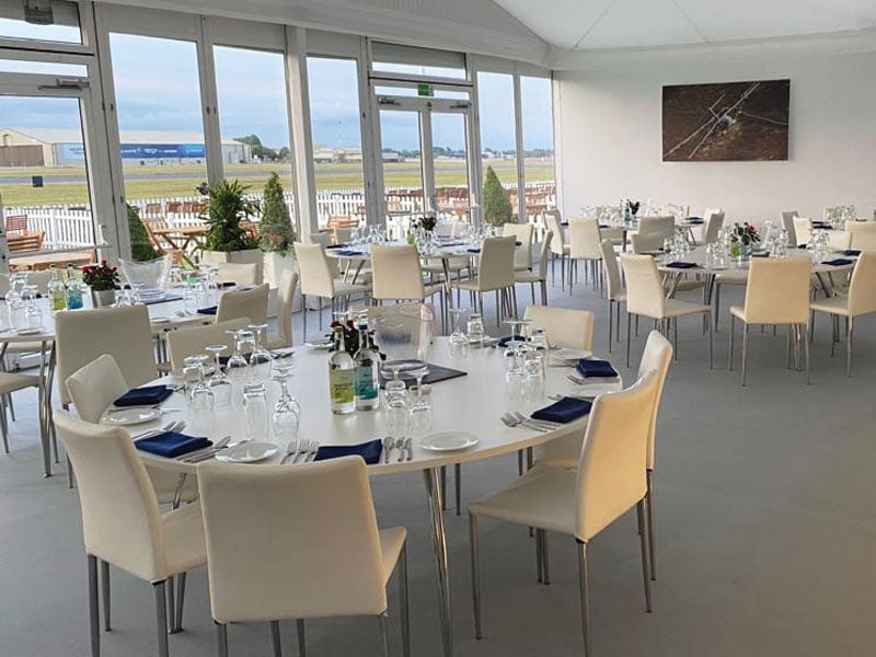 Bright dining room with round tables set with glassware, crockery and blue napkins, surrounded by cream chairs and overlooking an airfield through large windows.