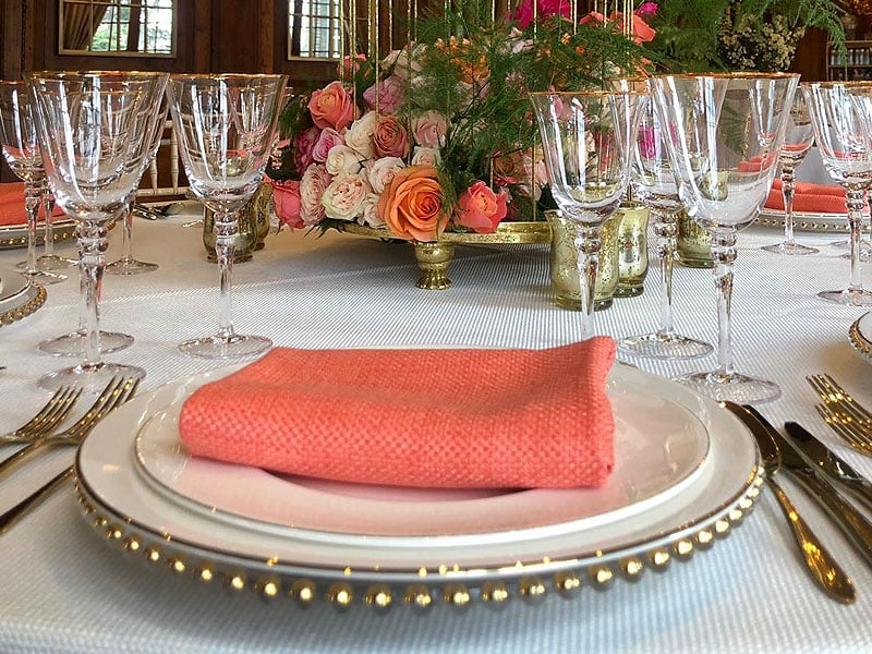 Formal dining place setting with gold-trimmed plates, a folded coral napkin, polished cutlery, crystal glasses and a floral centrepiece of pink and orange roses.